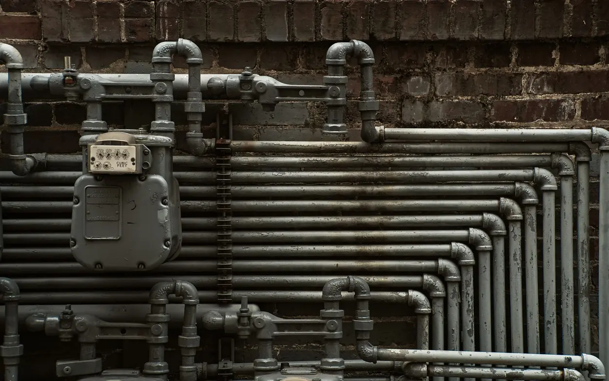 Rows of galvanized gas piping and an old dial gas meter mounted against a weathered brick wall