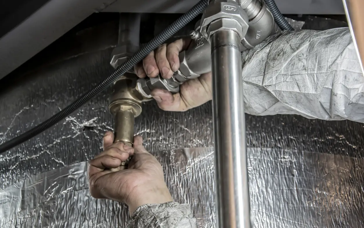 Close view of a plumber's bare hands holding a wrench on a brass valve fitting below a foil-wrapped duct